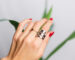 Woman hand with red manicure and two rings on fingers, on beautiful green palm leaf tropical background. Gray wall behind.